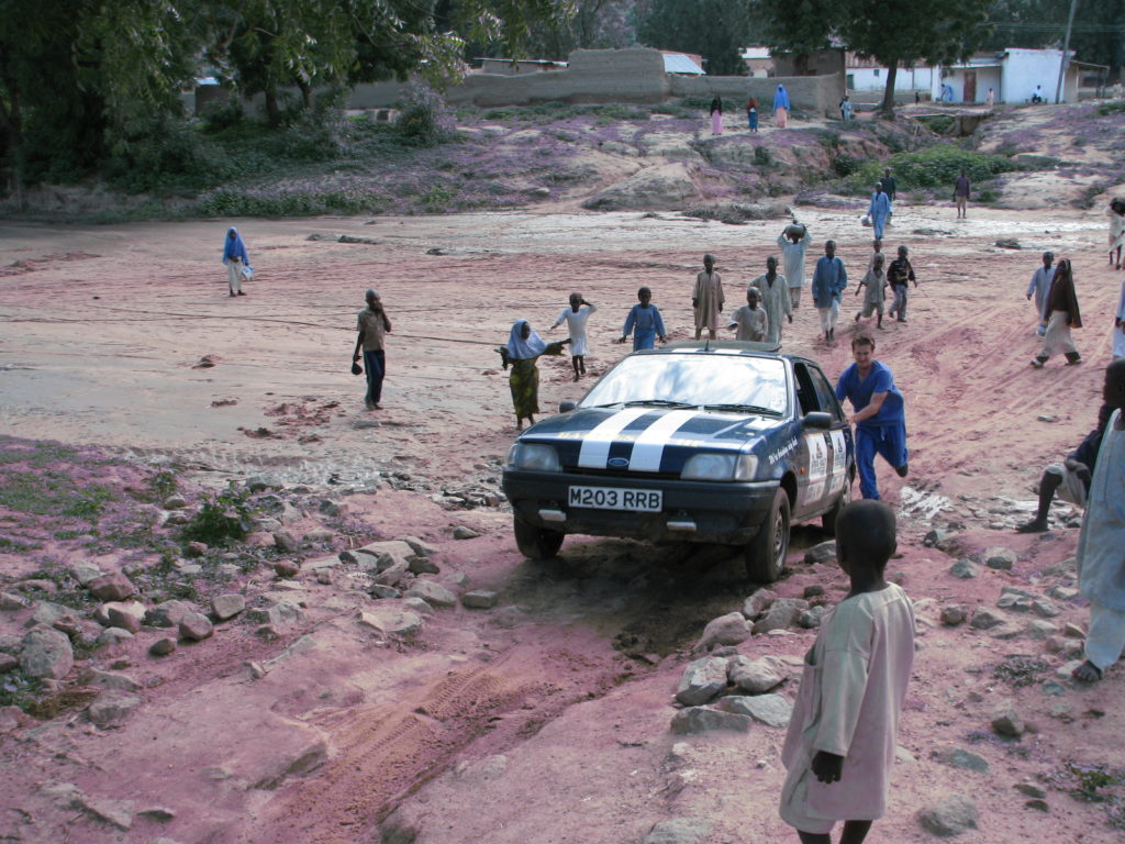 A Ford Fiesta being pushed along a rocky, dirt track while lots of people look on from the side of the road.