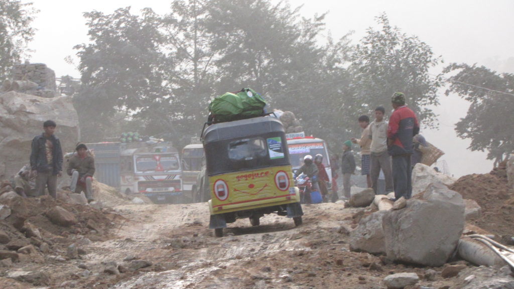 Joanne's yellow rickshaw drives along a rocky, dirt track with people watching from the side of the road.