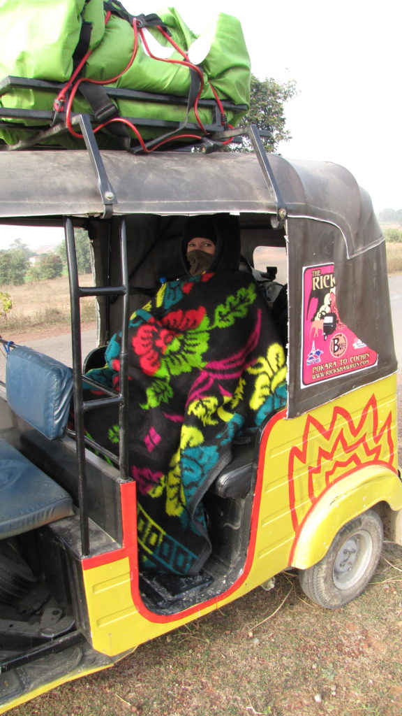 Joanne in a tuk-tuk/rickshaw. The rickshaw is yellow and Joanne is wrapped in a colourful blanket.