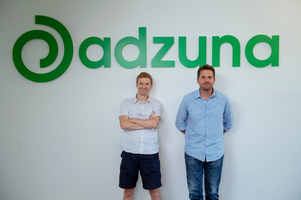 Doug and his Adzuna Co-Founder standing in front of a green sign reading 'adzuna'.