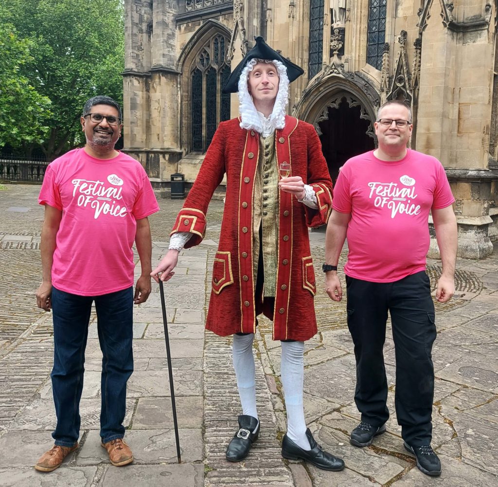 Samir Savant in a pink 'Festival of Voice' T-shirt. He is standing next to a man dressed as Handel and another man also in a pink 'Festival of Voice' T-shirt.