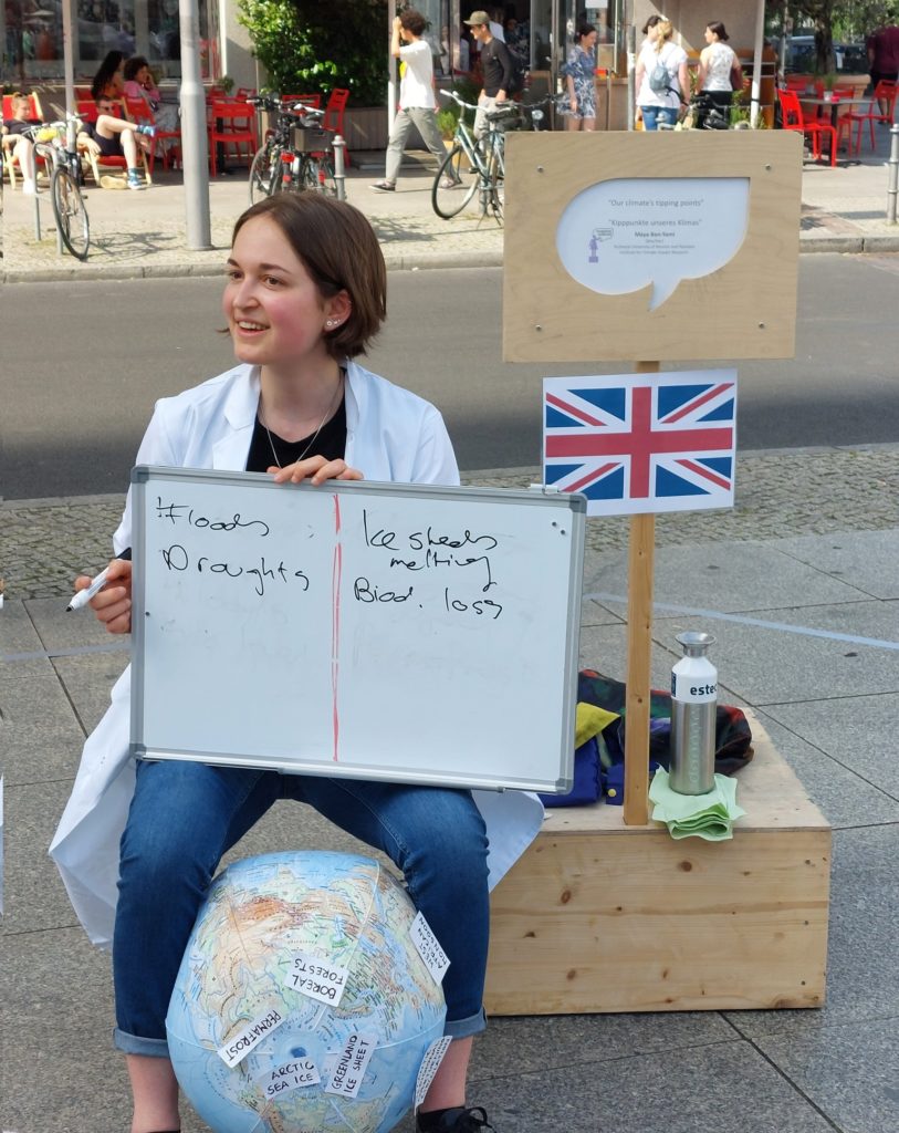 Maya at Soapbox Science Berlin. She is holding a small whiteboard with climate change consequences on it and sitting on an inflatable globe.