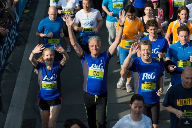 This is an image of Katharine running (presumably a marathon). Katharine is wearing a blue Proctor & Gamble running vest. She has her hands in the air in a wave and is smiling. She is next to some other runners, some of whom are also wearing P&G vests.