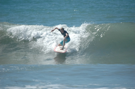 Katharine is surfing on a large wave. She is wearing a black top and blue board shorts.