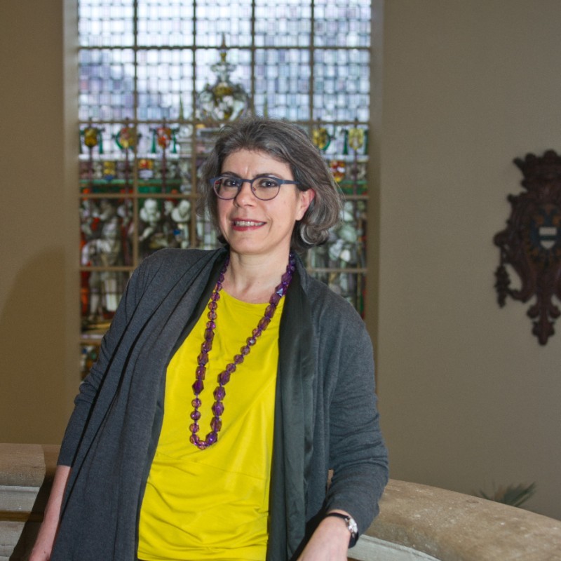 Photograph of Sofia Voutsaki. She is standing in front of a stained glass window wearing a yellow shirt, large purple beaded necklace and a grey cardigan. She has a dark grey bob haircut and wears glasses.