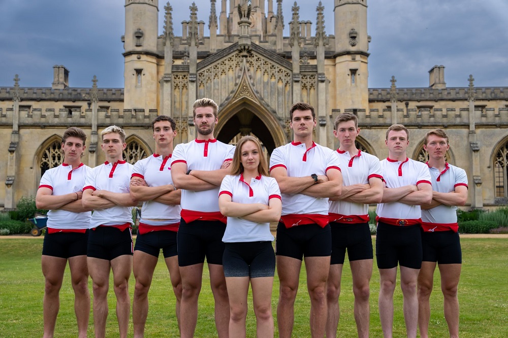 The eight rowers from the M1 squad stand behind their cox in front of New Court. They are all wearing black shorts and white polo shirts with red piping around the collar and short sleeves.