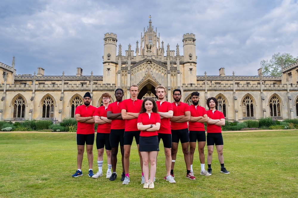 The eight rowers from the M3 squad stand behind their cox in front of New Court. They are all wearing black shorts and red polo shirts with white piping around the collar and short sleeves.