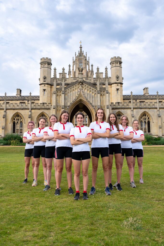 The eight rowers from the W1 squad stand behind their cox in front of New Court. They are all wearing black shorts and white polo shirts with red piping around the collar and short sleeves.