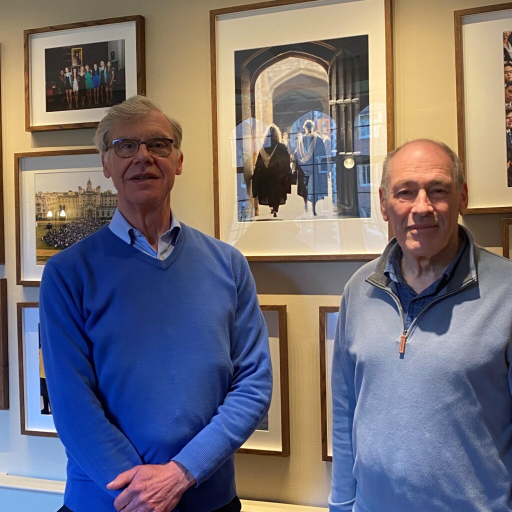 Mike Smith and Peter Stickland are both wearing blue jumpers. They are standing in front of some photos of College life in the new SJC bar.