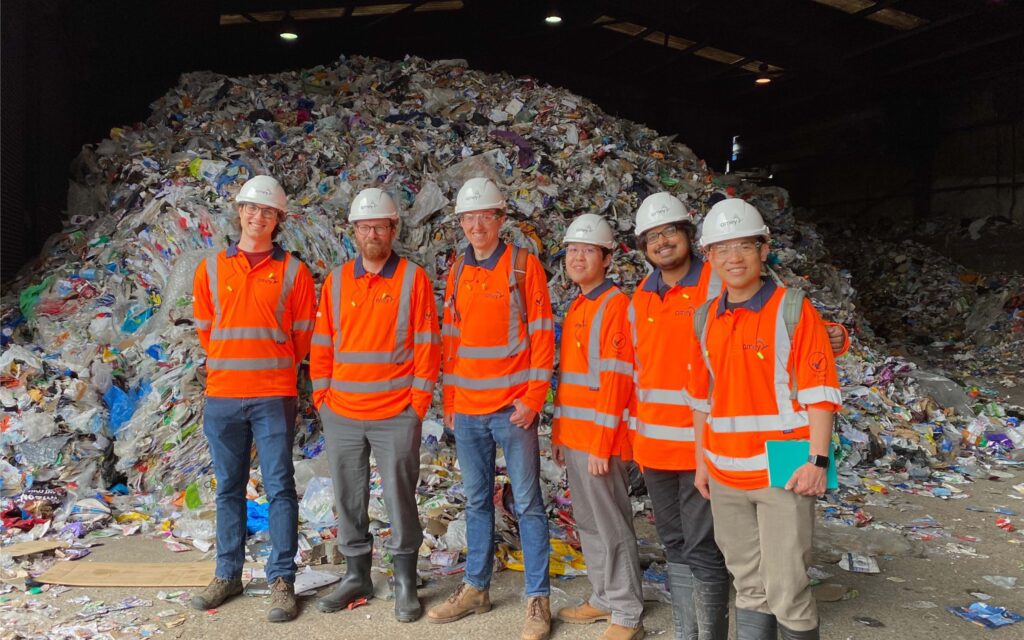Erwin and colleagues at Amey recycling plant. They are wearing orange hi-vis jackets, white plastic helmets and standing in front of a heap of recycling.