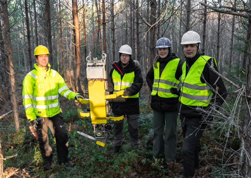 Three men and Caroline are standing in a wood with their Airforestry tool. All of them are wearing high vis vests and hard hats.