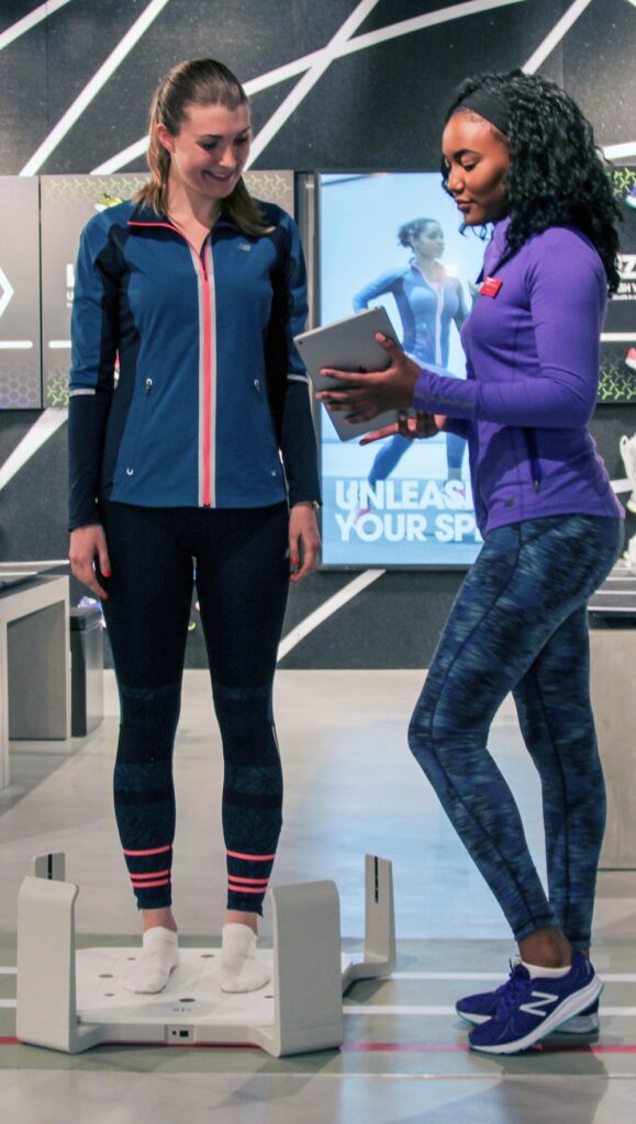 Caroline is wearing fitness gear and standing on the Volumental 3D foot scanner in a running shop. A shop assistant is holding an IPad and showing it to Caroline. 