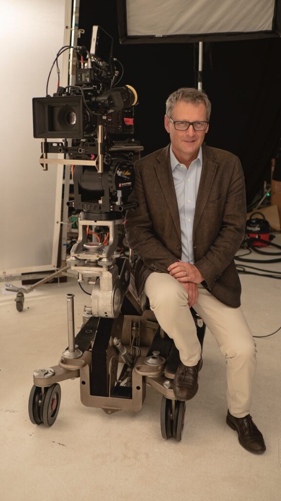 Colin is sitting at the base of a large camera unit. He is dressed smartly in a dark blazer with light chinos. He is sporting glasses and a smile.