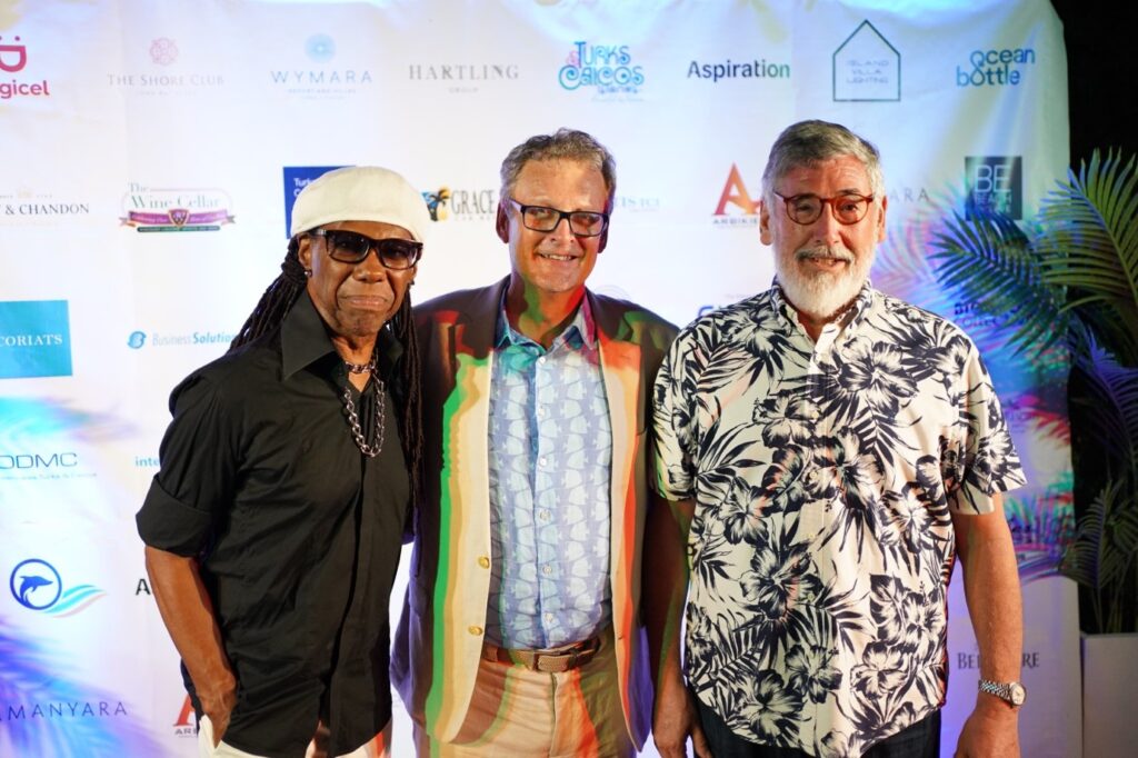 Colin has his arms round musician Nile Rodgers and director John Landis. Colin is wearing a smart shirt with a 'fishy' pattern and a blazer. Nile has a black shirt with rolled up sleeves, a light hat, shades and beads around his neck. John is in a black and white hawiian shirt.