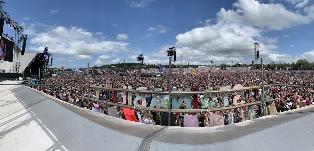A behind-the-scenes shot from on a stage at Glastonbury. Thousands of people can be seen behind the 'GLASTONBURY' lettering which is backwards from this angle.