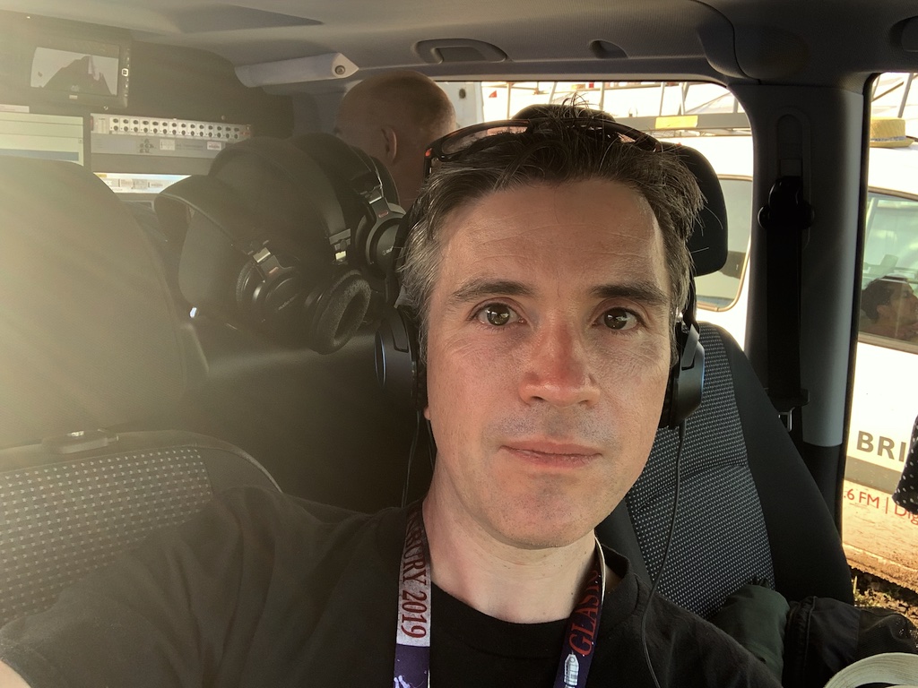 Mark is in a radio van. He is wearing headphones and a lanyard. Behind him at van seats with more headphones on.