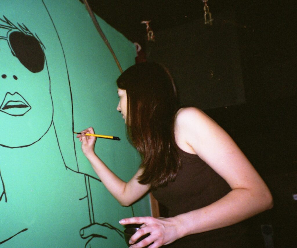 Imogen is up a ladder painting an illustration of a woman's face on a green wall.