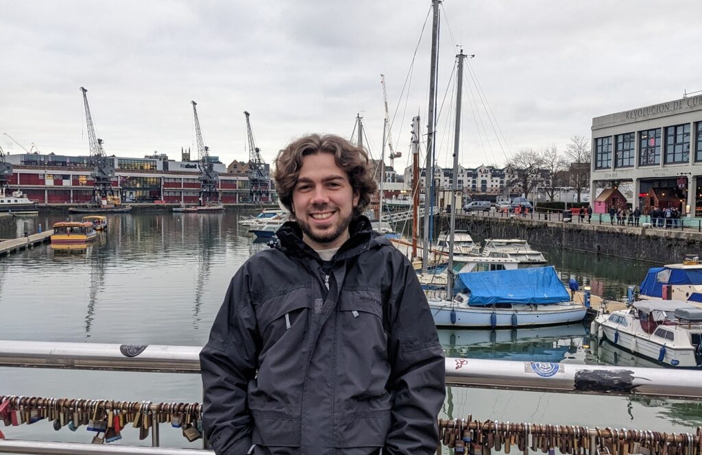 Jamie is wearing a black raincoat and is standing in front of a busy marina. He has dark curly hair and is smilling. Padlocks are attached to the railings behind him and there are boats on the water behind.