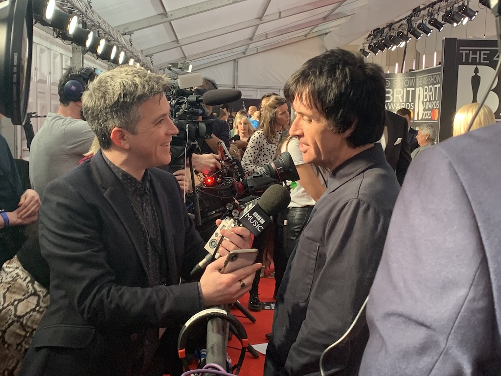 Mark is standing next to Johnny Marr. He holds a BBC Music microphone. They are both smiling.