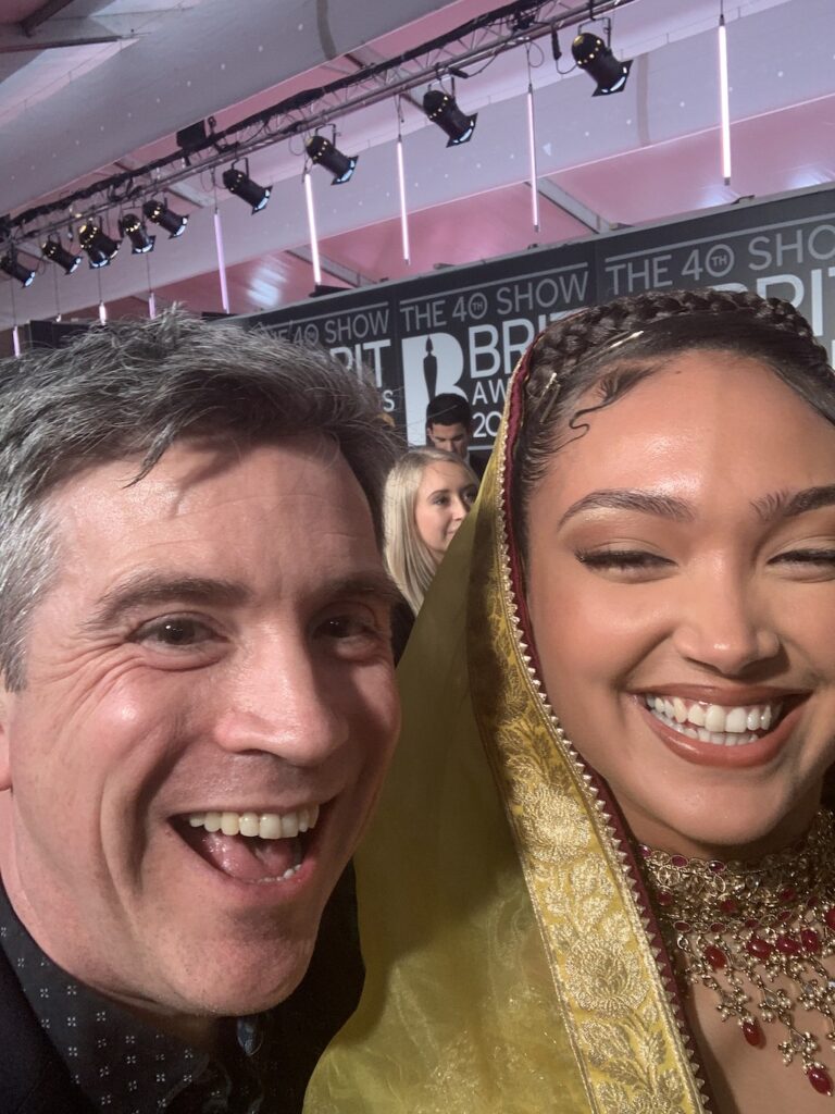 A selfie image of Mark and Joy. Both are smiling. Joy has a gold coloured headscarf and an elaborate red and gold necklace.