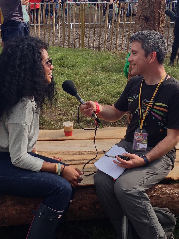Mark and Kelis sit opposite each other on a bench. Mark is pointing a microphone at her. Both are smiling.