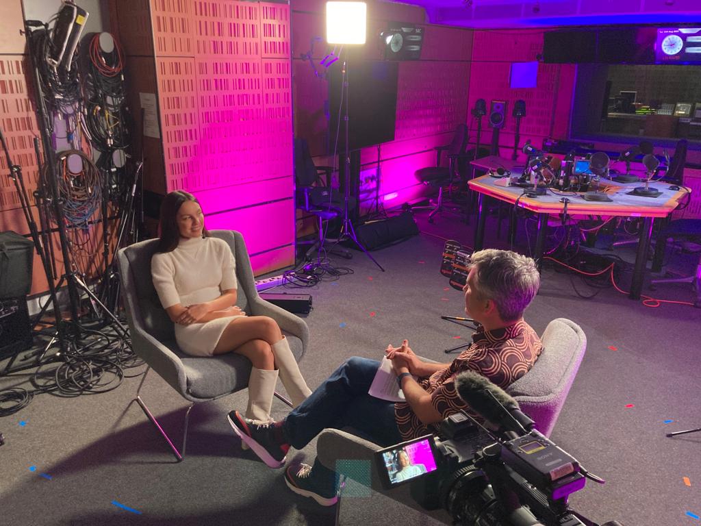 A shot from inside a studio. Mark and Oliva are sitting in arm chairs opposite one another. The room is uplit in pink and the camera points at Olivia