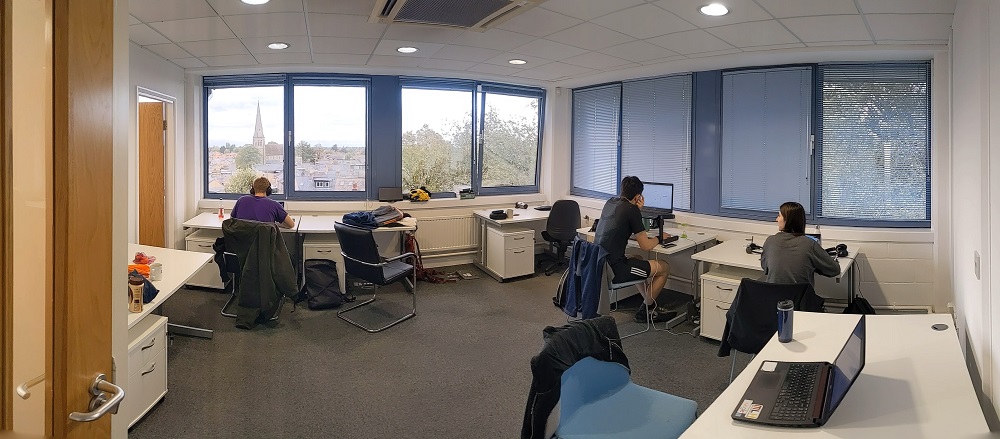 Three workers are seated at desks. Outside the window there is a view of the Cambridge skyline.