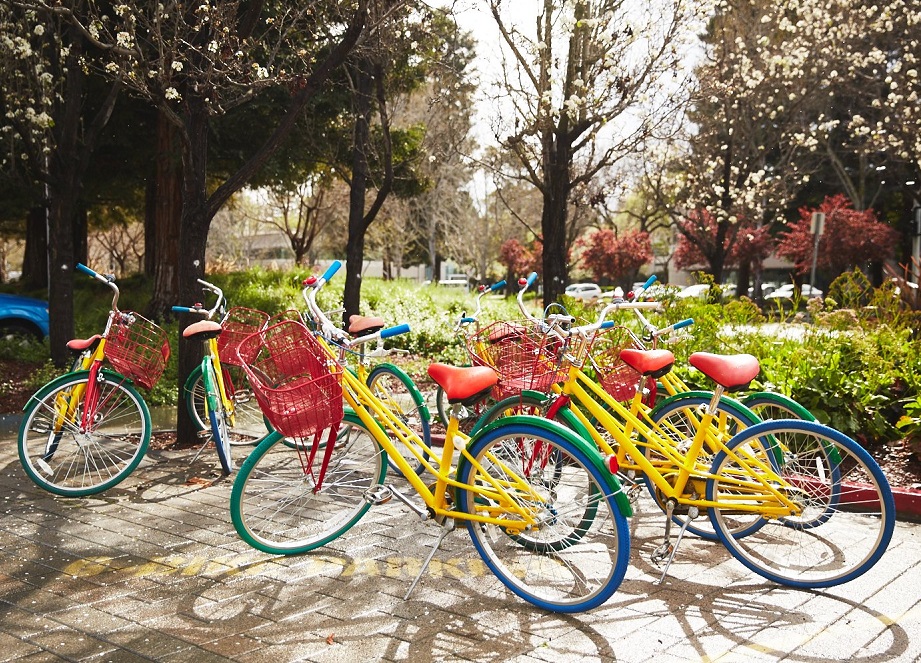 Brightly coloured bicycles of yellow, green, blue and red. They are parked by a green park with trees.
