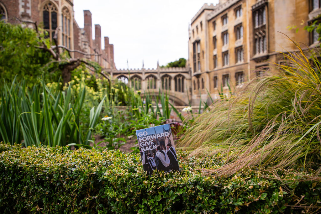 Go Forward Give Back brochure is balanced on a hedge in the Master's garden. Behind it are some flowers and the Bridge of Sighs. 