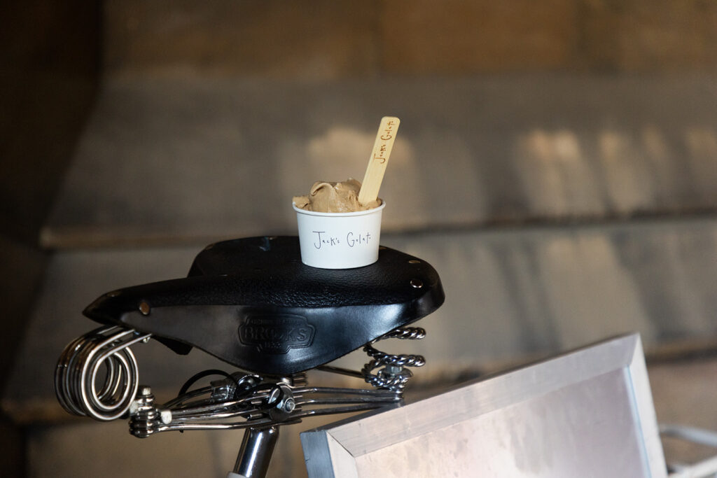 A bike seat with a pot of ice cream on it. The ice cream pot says 'Jack's Gelato' on it and a wooden spoon sticks out of the top. It looks like a chocolate ice cream.