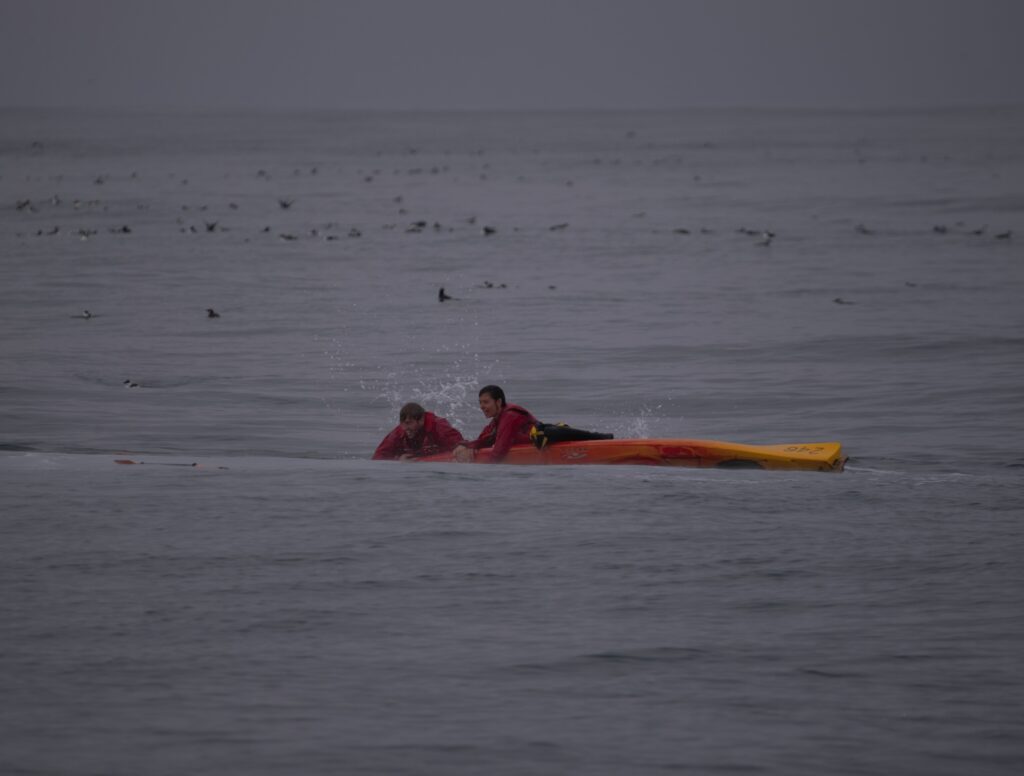 Tom and Charlotte cling to their kayak in the sea. The kayak is orange.