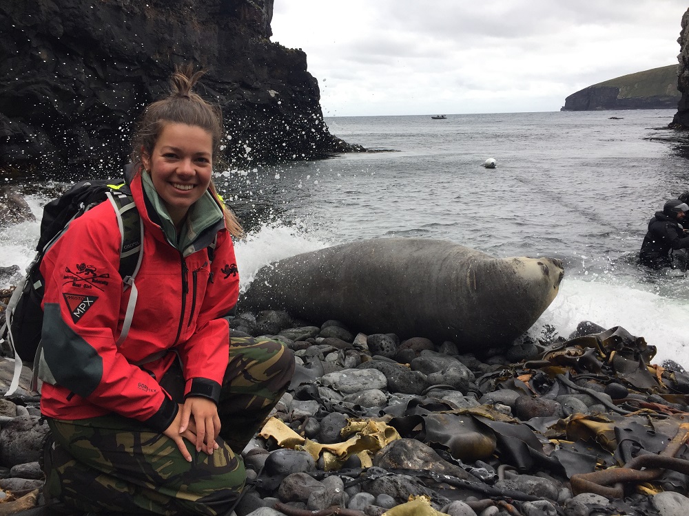 Kayla is kneeling down on a pebbly beach next to a seal. The sea spray surrounds them and there are cliffs in the background.