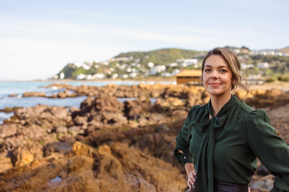 Kayla is standing at the right of the image. She is standing on a rocky beach with green hills in the background. Kayla is wearing a dark green blouse with a bow at the neck.