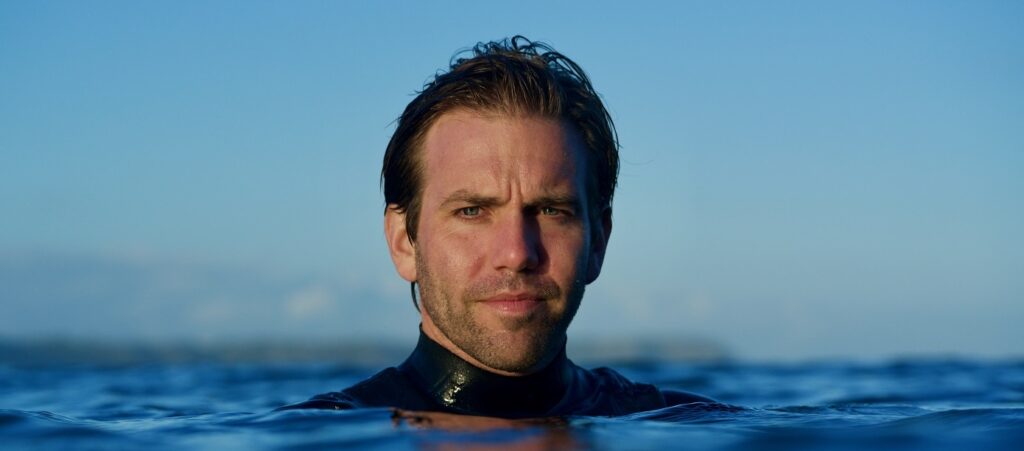 A headshot of Tom. Tom is wearing a wetsuit and only his head is visible above the sea's surface. He has dark blond hair, blue eyes and stubble.