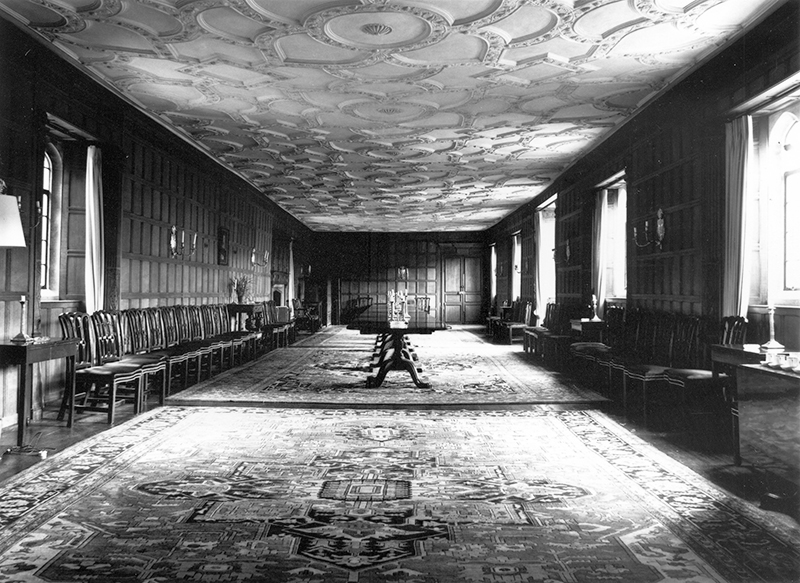 The Senior Combination room is wood-panelled and has three large rugs on the floor. A table is in the middle and seats are against the walls.