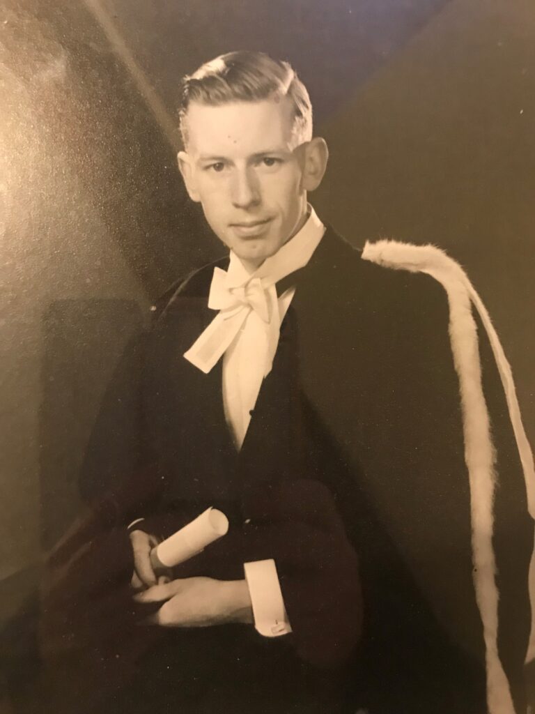 A young Donald Rushton poses with a gown and scroll for graduation. The image is black and white.