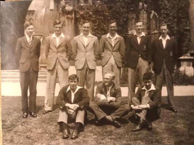Six men in suits stand in front of a College building. Three men are seated on the ground below them. The photograph is in black and white.