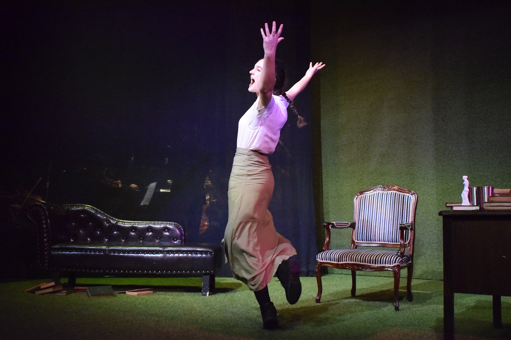 A woman with plaits raises her hands in the air. One of her feet is lifted off the ground. Household furniture is behind her.