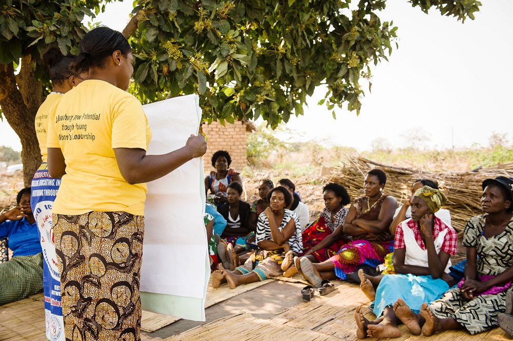 Two women in yellow t-shirts hold some large flipboard paper and are presenting to women seated on the floor. They are outside under a tree