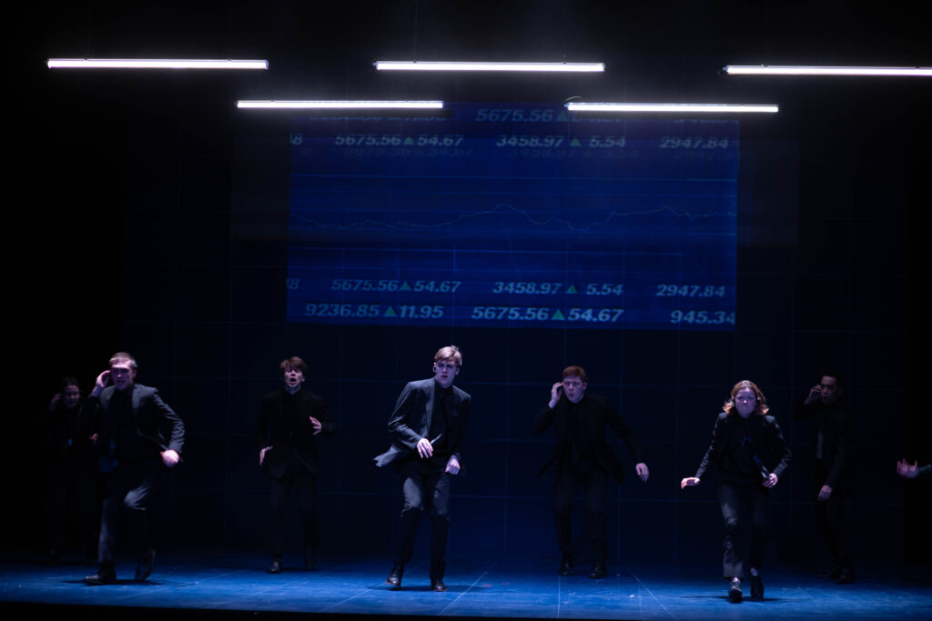 Smartly dressed cast members standing spaced apart on stage. Numbers are projected on a screen behind them.