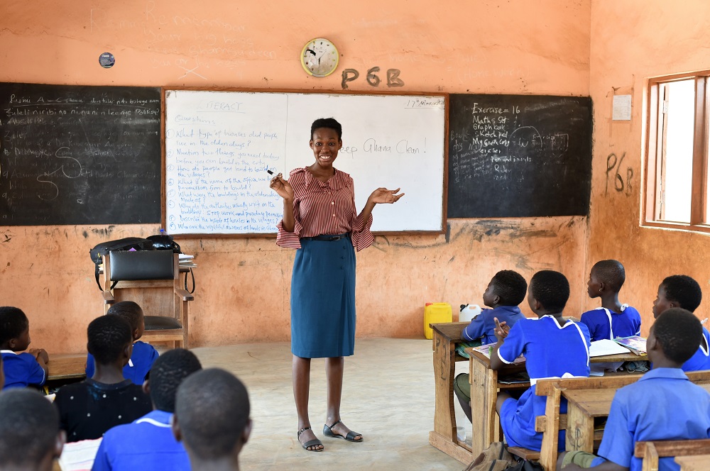 A female teacher stands in front of whiteboards and chalk boards teaching children in blue uniforms