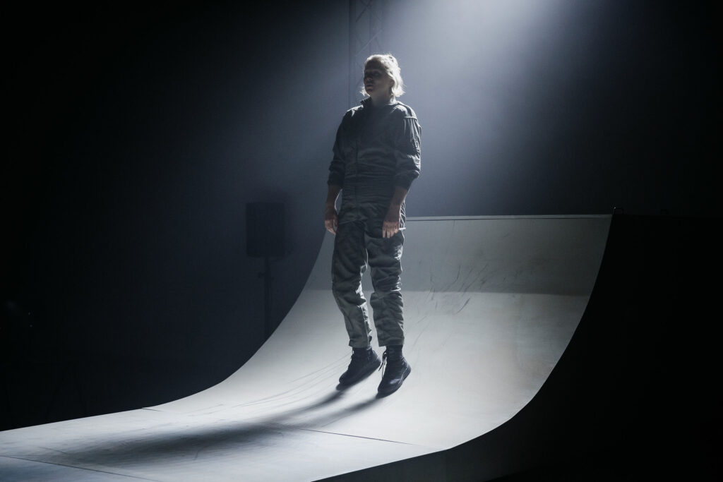 A woman in a boilersuit stands half way up a skating ramp