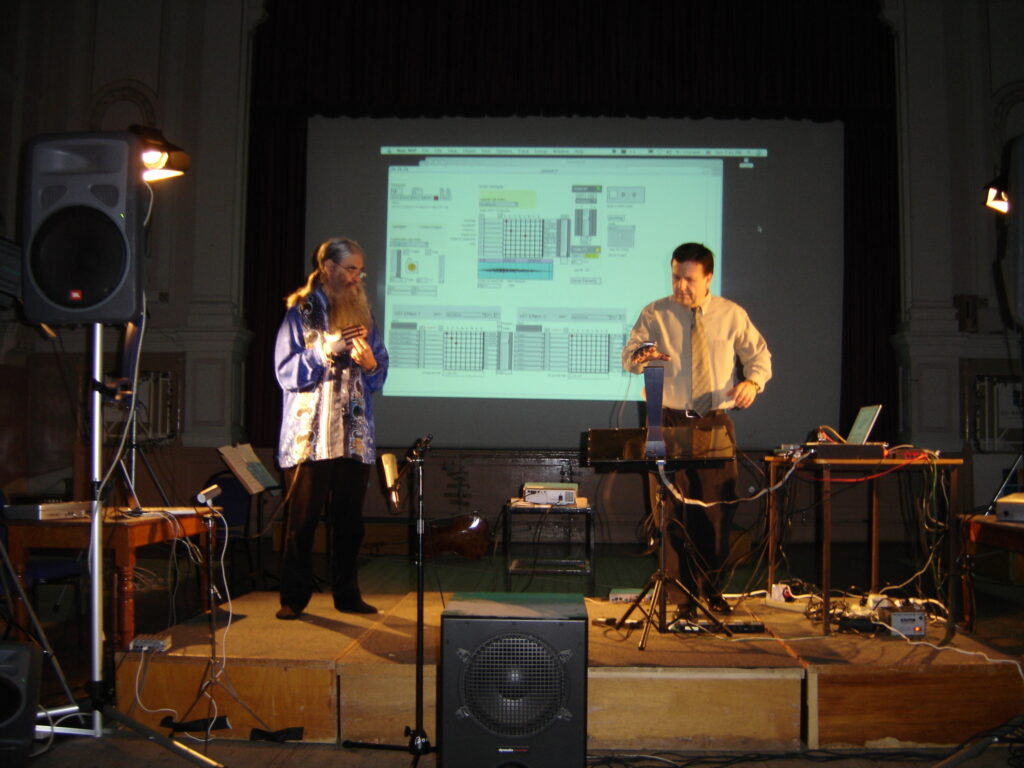 John and Dr B stand in front of a projector screen with their instruments on a stage