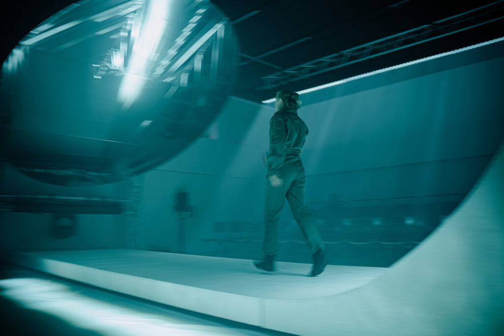 A woman in a boiler suit strides down something that looks like a skatepark structure. 