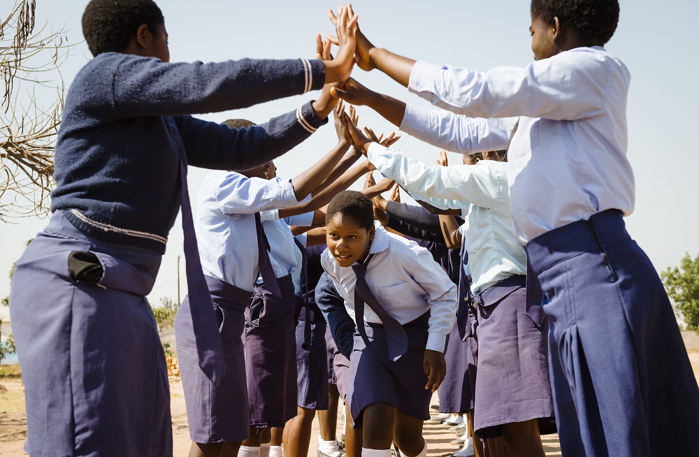Female students make arches in pair and another girls bends down to walk through it. They are wearing blue school uniforms