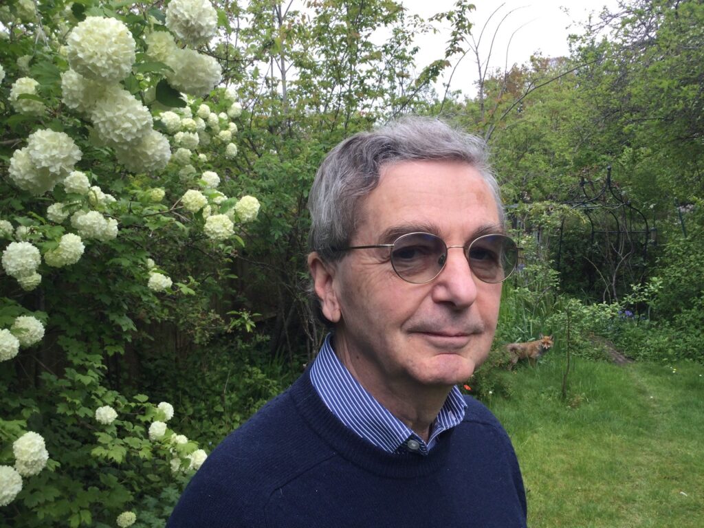 Kieth is standing in a typical English garden. He is wearing a blue collared shirt and blue jumper and dark glasses. He has short grey hair. Behind him there is a fox.