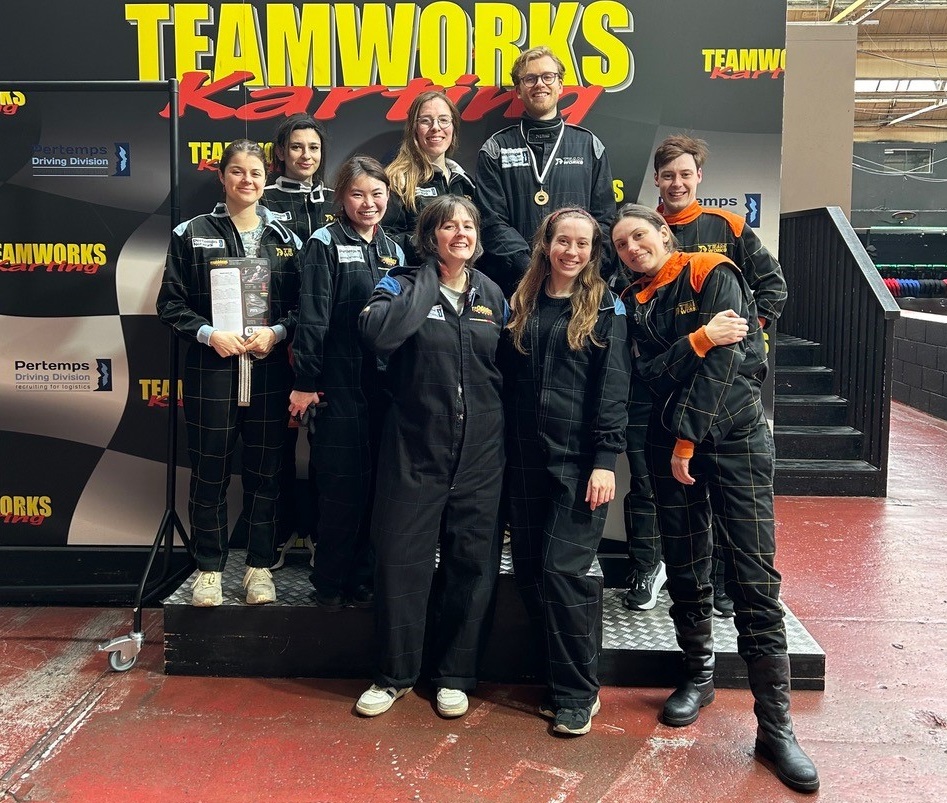 Amy and eight members of her lab group are dressed in black-overalls in front of a sign reading 'Teamworks karting'.