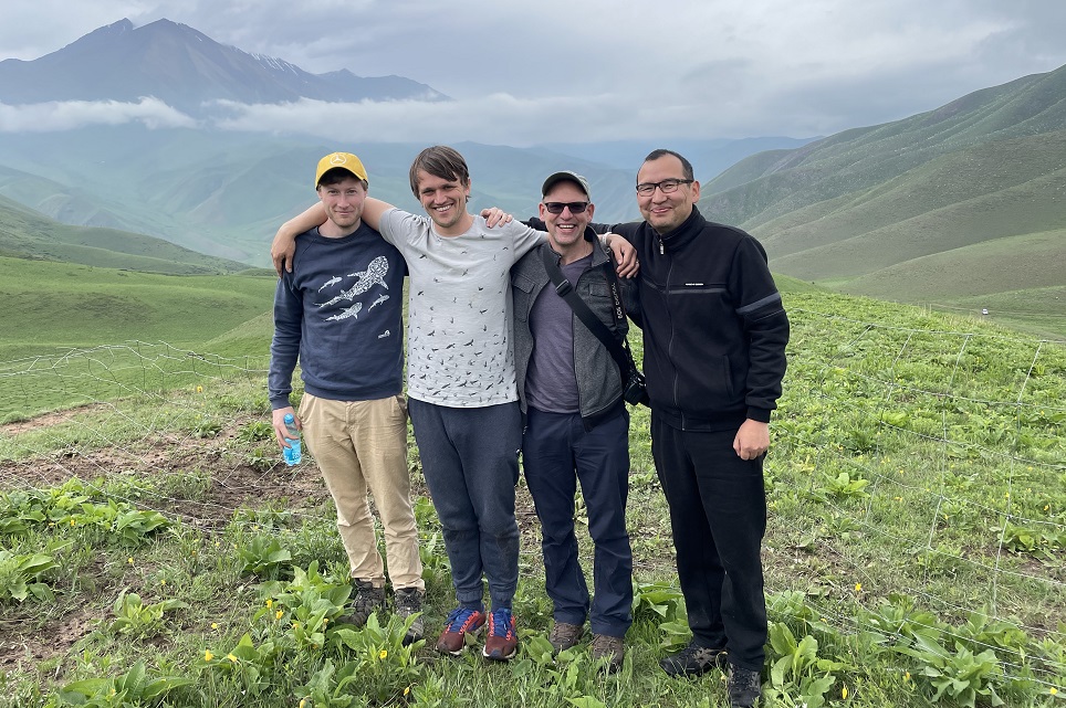 Brett Wilson, David Gill, Maarten Christenhusz and Ormon Sultangaziev stand arm in arm smiling. Behind them are mountains and clouds.