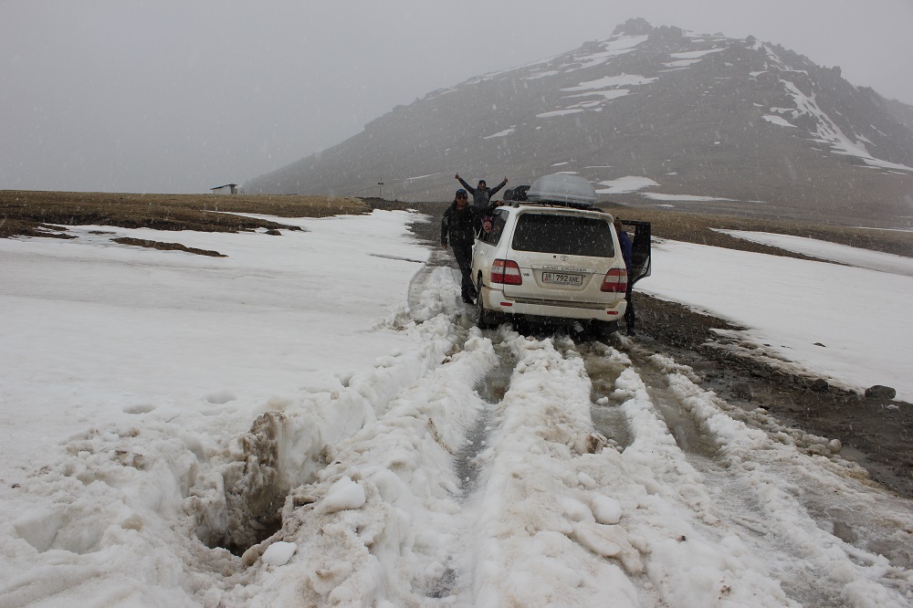 The team's car is making tracks through deep snow. Members of the team hang out of the windows and doors. In the background is a mountain and it is snowing.