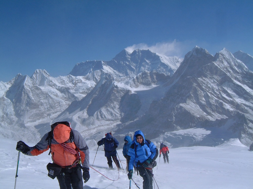 A group of hikers in warm hooded coats, with ropes and poles trek through the snow. Mountains peaks are behind them. 